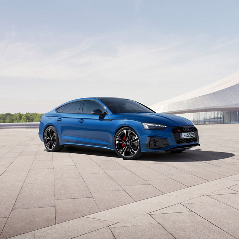 Blue Audi sedan parked on an open plaza with a clear sky and modern architecture in the background.