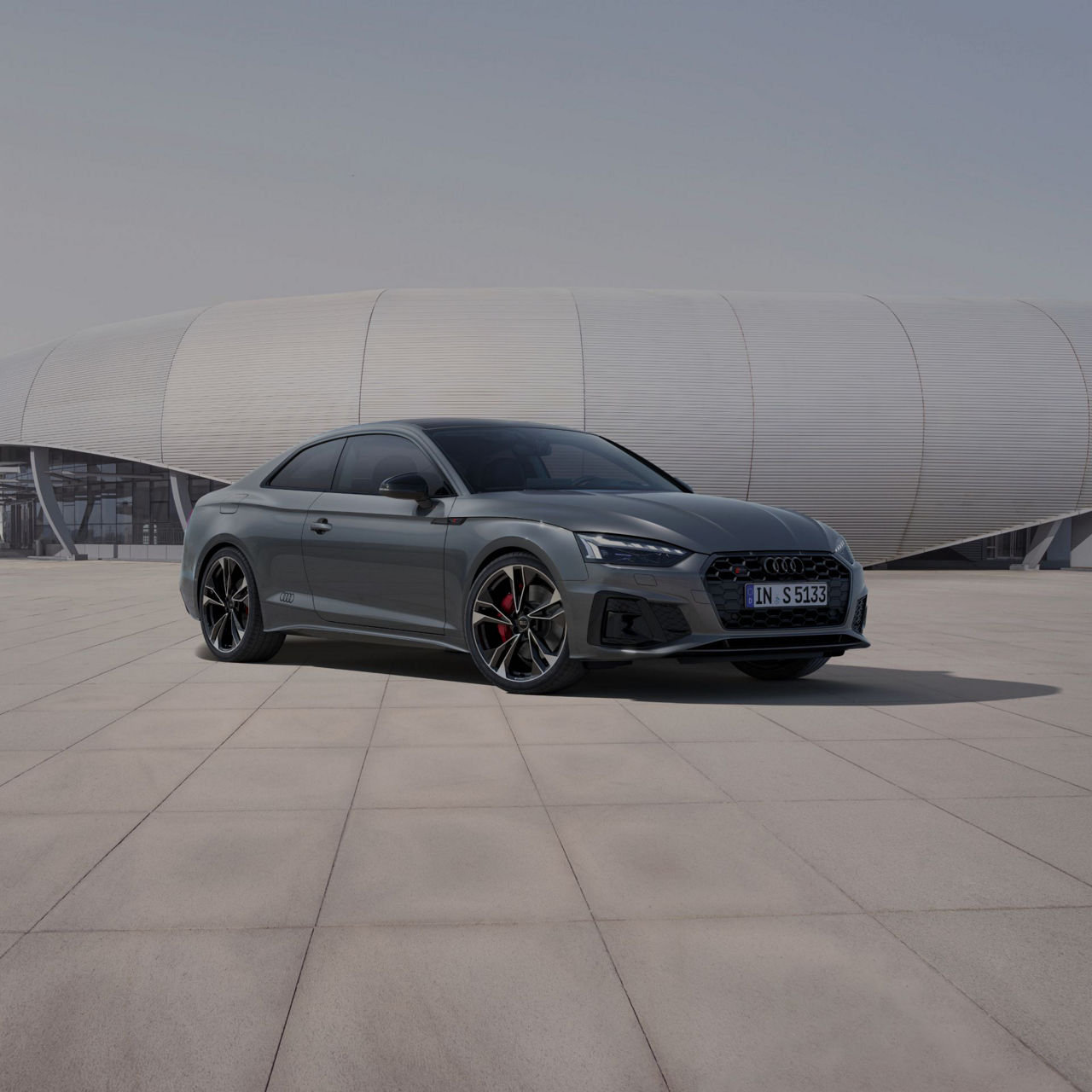 A grey Audi S5 coupe parked in front of a futuristic building with a curved facade.