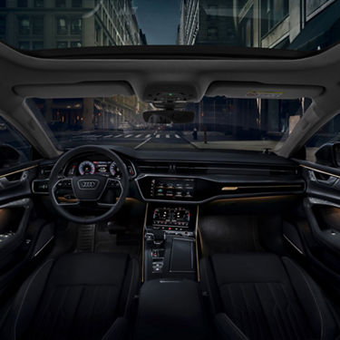 Interior view of a car with steering wheel and dashboard, city street visible through windshield.