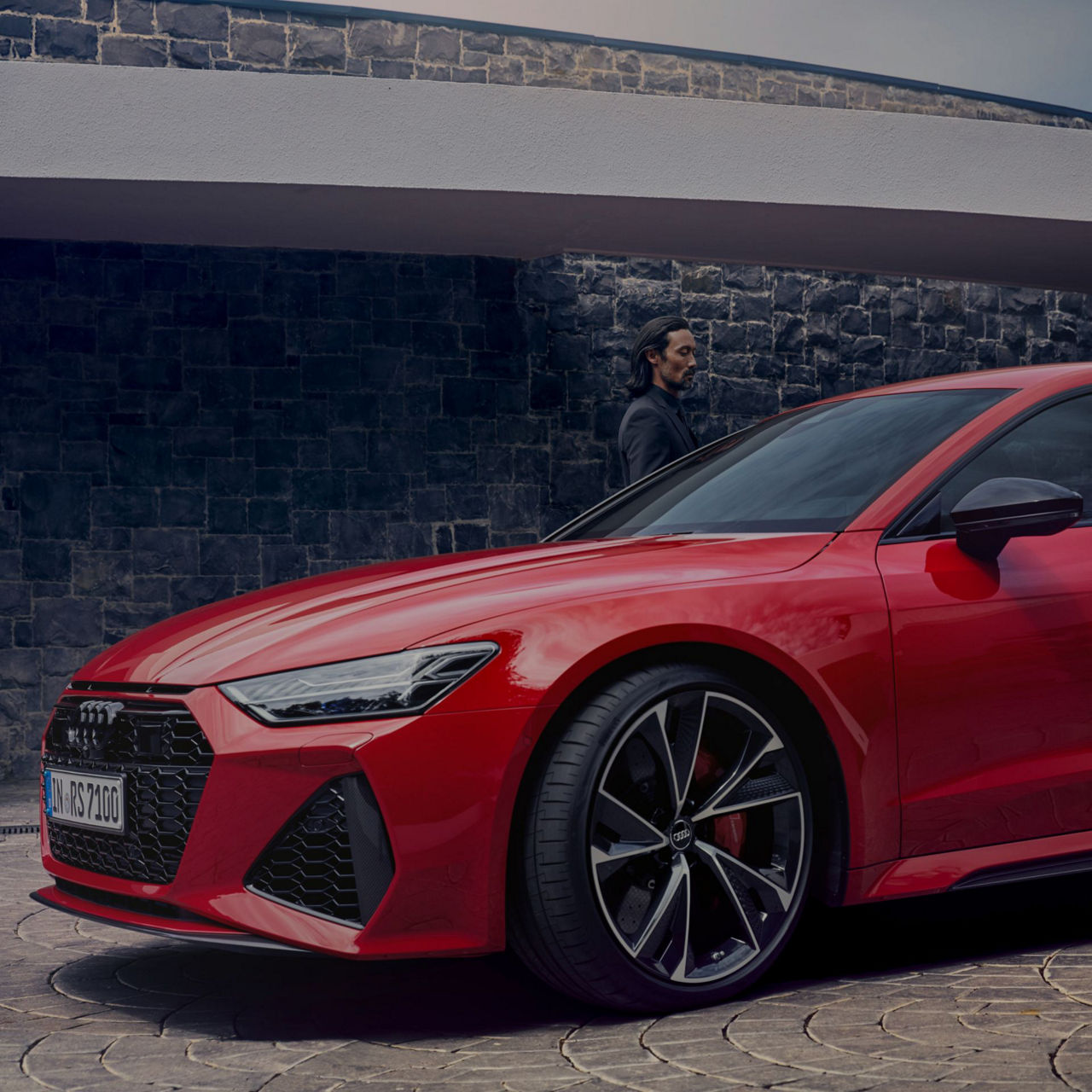 Red car parked with a person standing behind it, against a stone wall under an overhang.