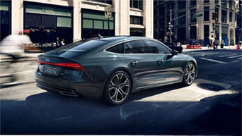 A grey Audi car parked on a city street with blurred traffic and pedestrians.