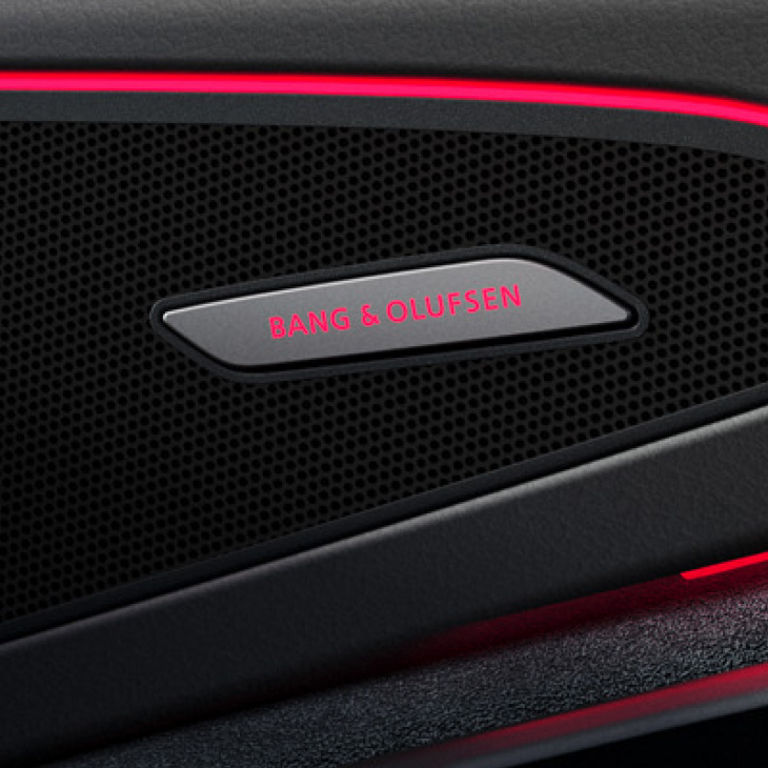 Close-up of a car speaker grille with 'BANG & OLUFSEN' logo and red ambient lighting.