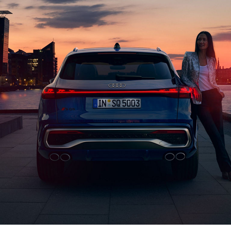 Blue Audi SQ5 parked by riverside at sunset with city skyline in background.