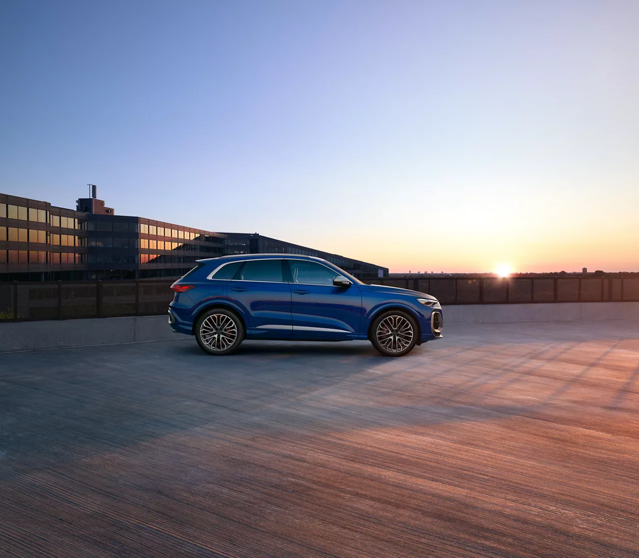 A blue SUV parked on a rooftop at sunset with buildings in the background.