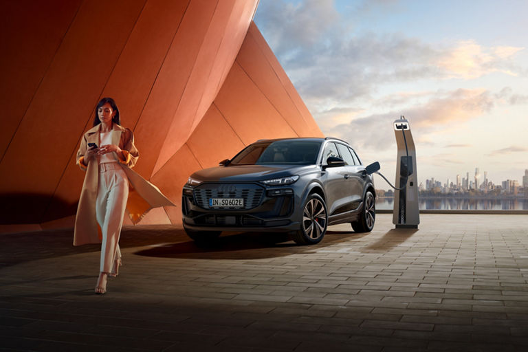 Audi Sq6 etron SUV charging at a station with a city skyline in the background at dusk.