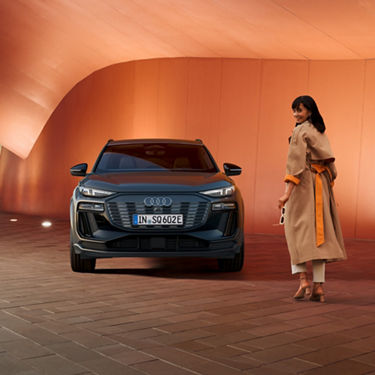 Woman in a beige coat standing next to a blue Audi in a modern architectural setting.