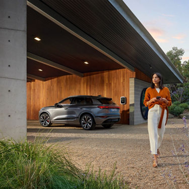 Audi SQ6 etron car parked under a carport attached to a wooden house, with a person standing in the foreground.