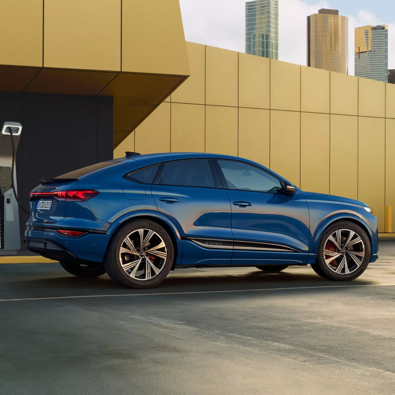 Audi SQ6 Sportback e-tron in a high walled yard near charging point with sky scrapers in the background.