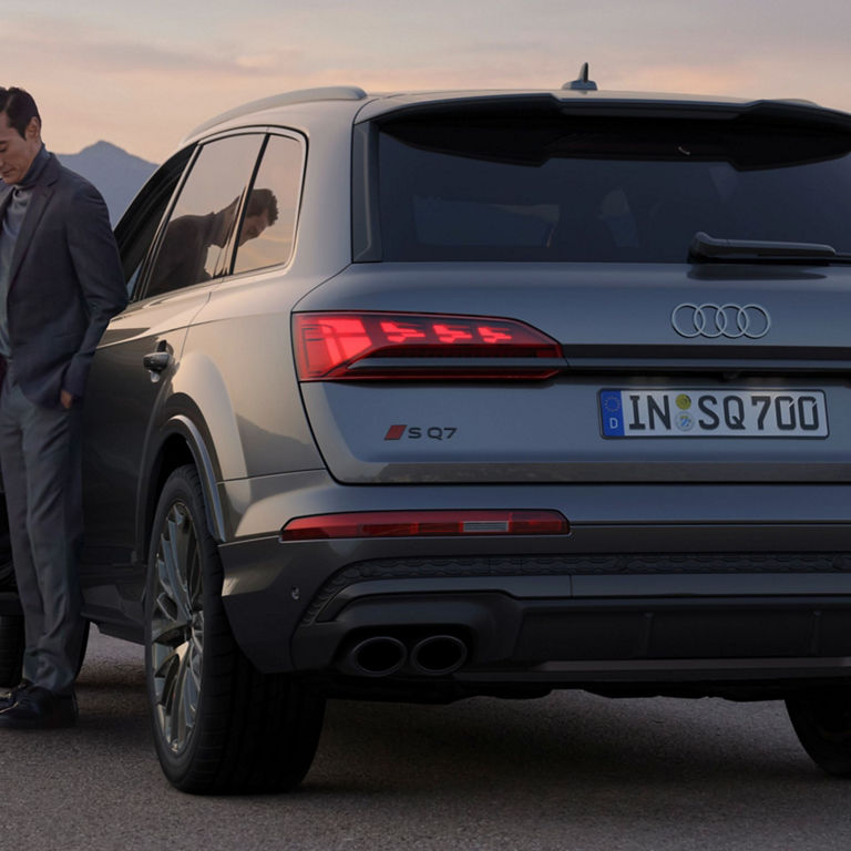 Person standing by an open door of a parked Audi SQ7 near the coast at dusk.