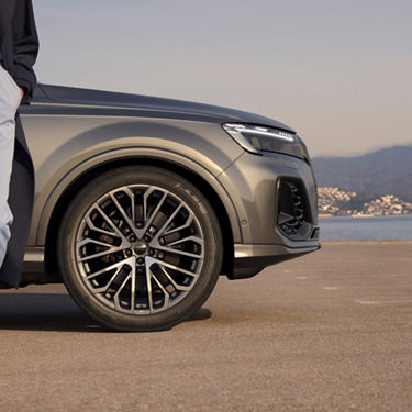 Silver SUV parked with close-up on the wheel, person standing beside it, overlooking a coastal town.