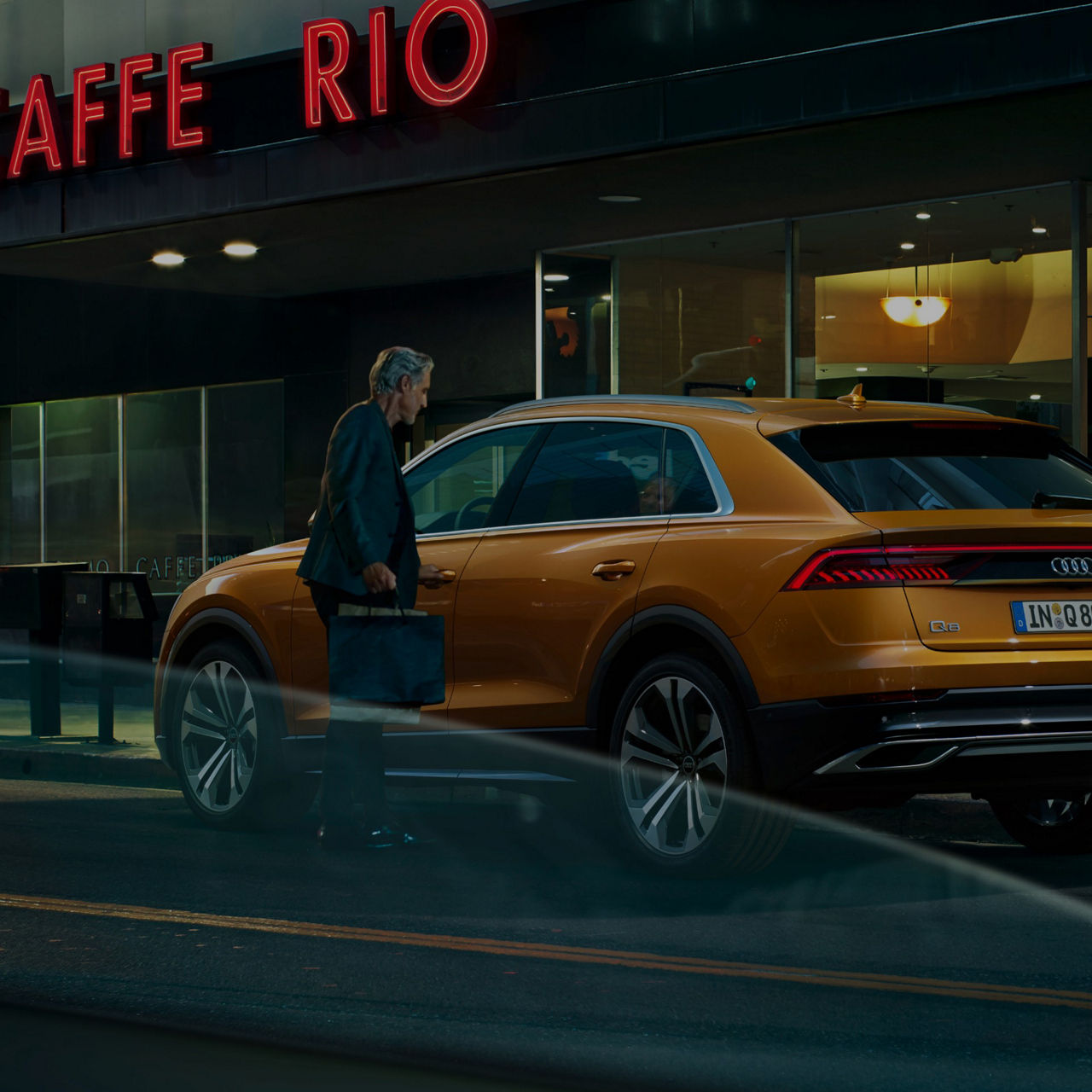 A person walking in front of a parked orange Audi with a lit "CAFFE RIO" sign in the background.