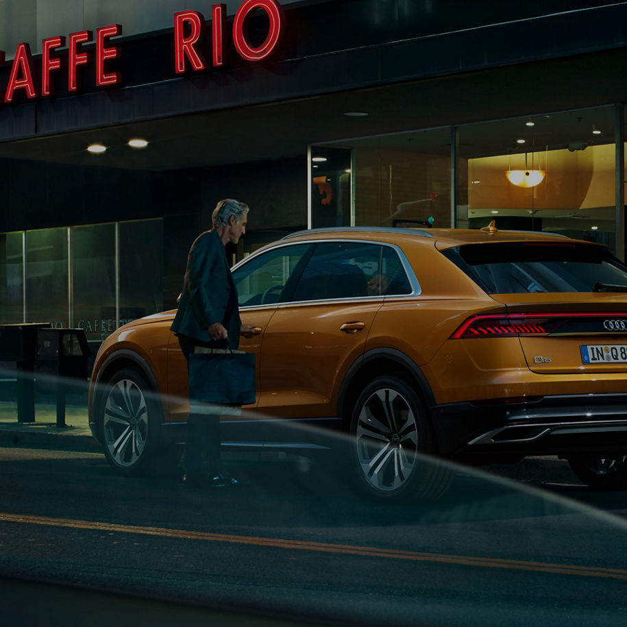 A person walking in front of a parked orange Audi with a lit "CAFFE RIO" sign in the background.