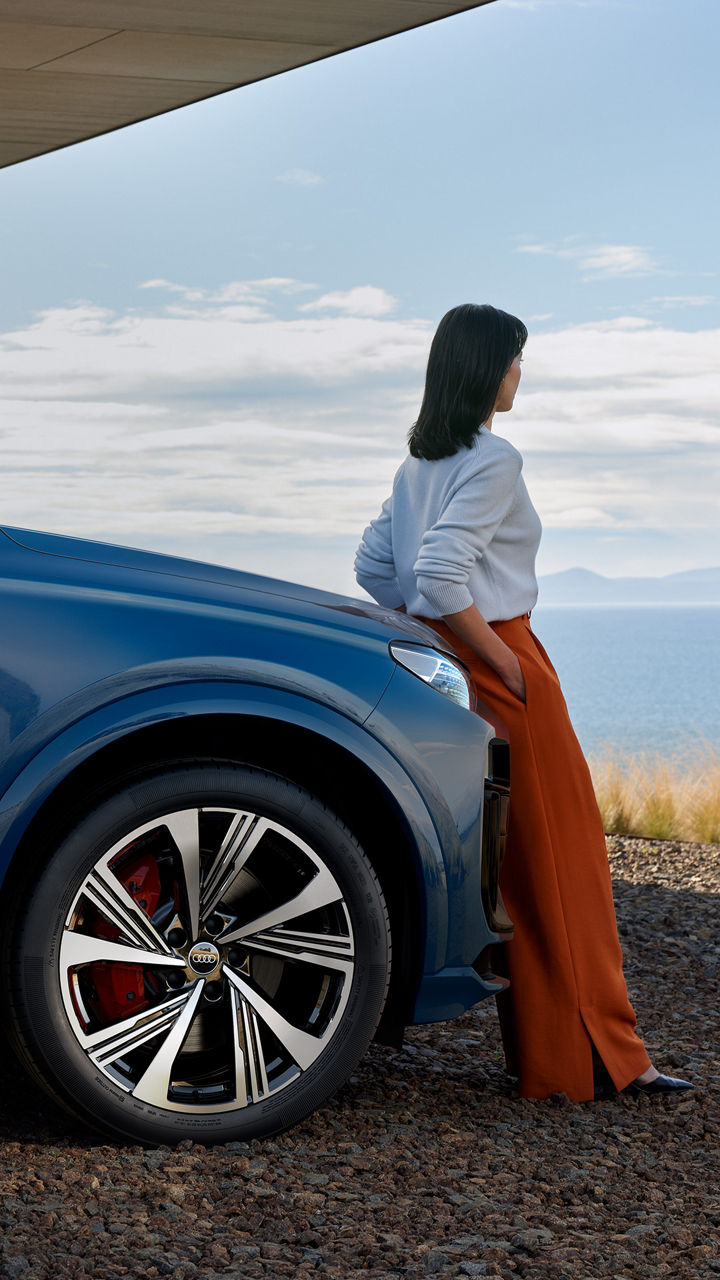Eine Frau steht an die Motorhaube eines blauen Audi-Modell gelehnt und blickt auf das Meer.