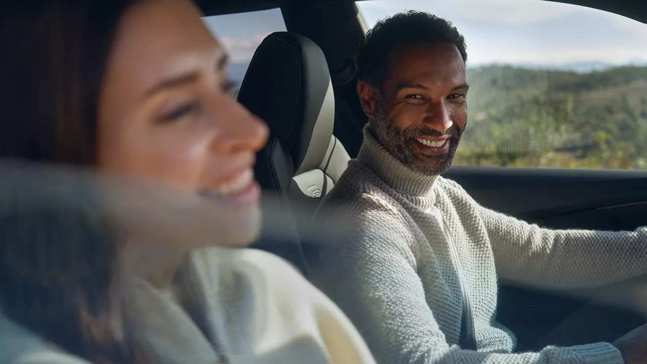 Eine Frau und ein Mann sitzen in einem Audi und lächeln sich an. Die Fensterscheibe auf der Beifahrerseite ist halb geöffnet.