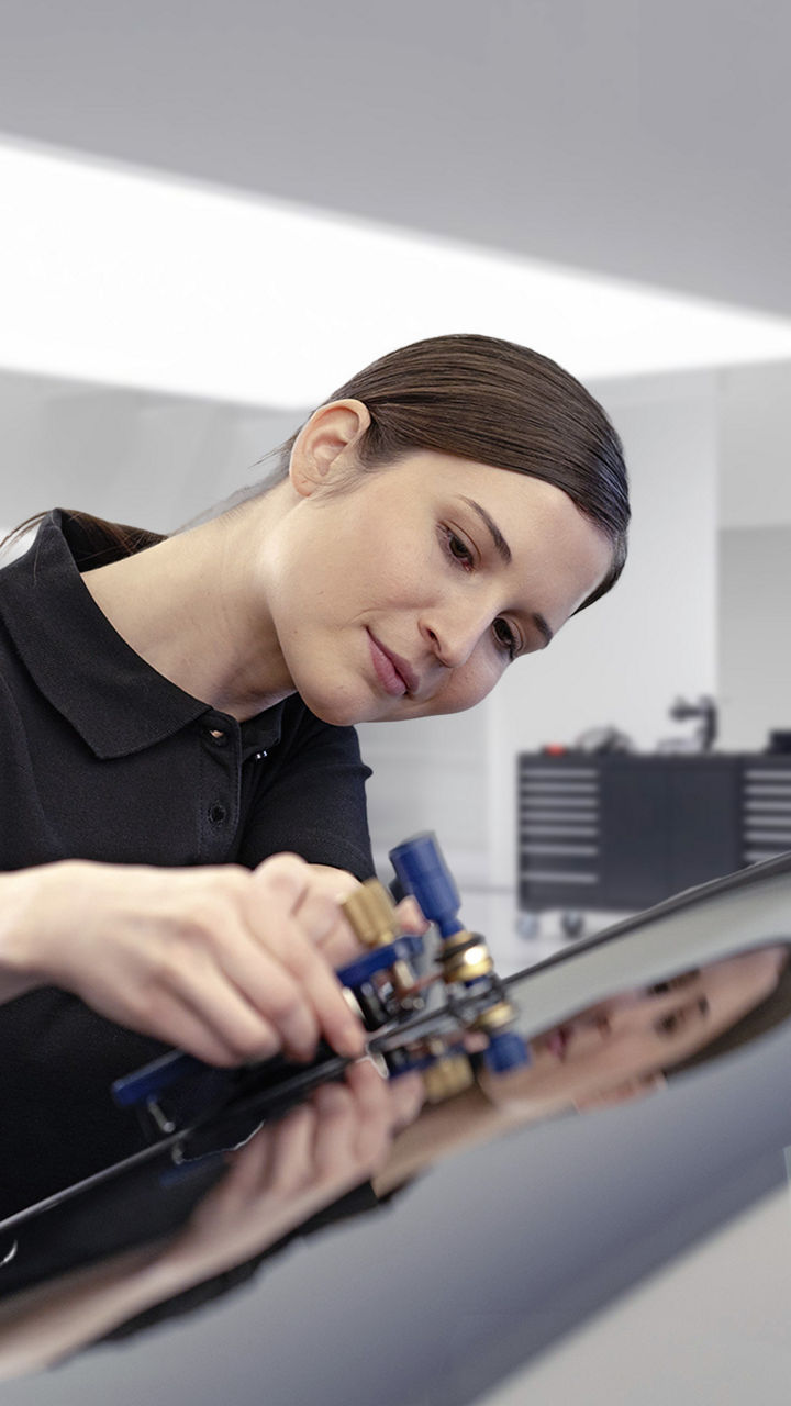 Eine Frau steht in einer Werkstatt und hält ein Gerät an eine WIndschutzscheibe eines Audis