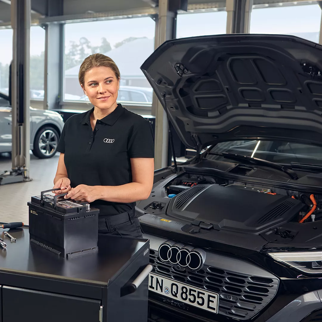 Eine Frau hält eine Audi Starterbattrie und steht vor einem Audi mit offener Motorhaube