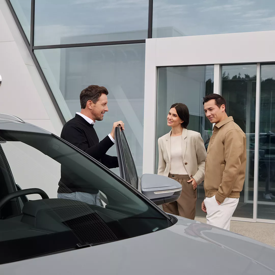 Drei Personen stehen vor einem Audi Autohaus und der linke Mann hält eine offene Auditür