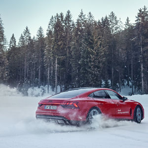 Roter Audi e-tron GT driftet auf schneebedeckter Fläche, wirbelt Schnee auf; Tannenwald im Hintergrund.