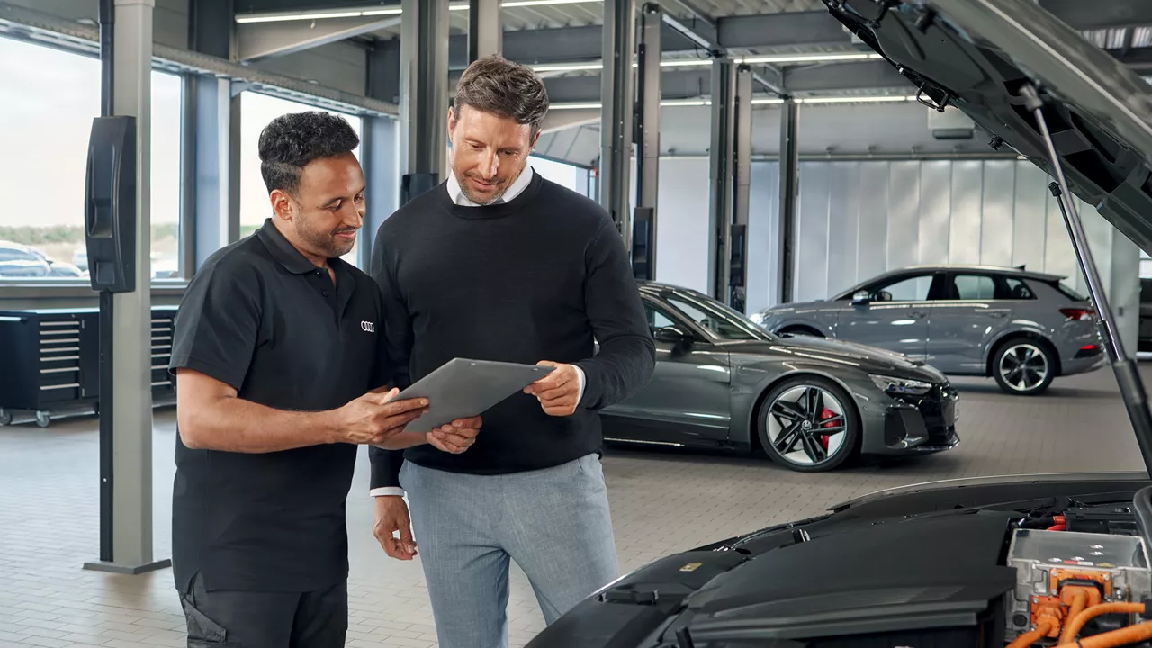 Zwei Männer stehen in einer Werkstatt vor einem Audi Modell mit offener Motorhaube und schauen gemeinsam auf den Screen eines Tablets.