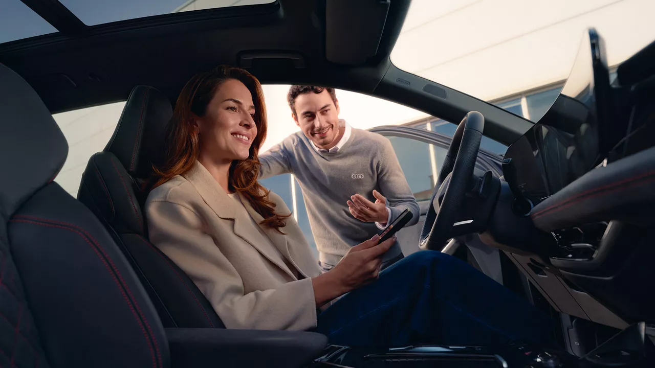 Frau sitzt lächelnd hinterm Steuer eines Audi Modell und hält ein Smartphone in der Hand, während ein Audi Mitarbeiter draußen am offenen Türrahmen steht und spricht.