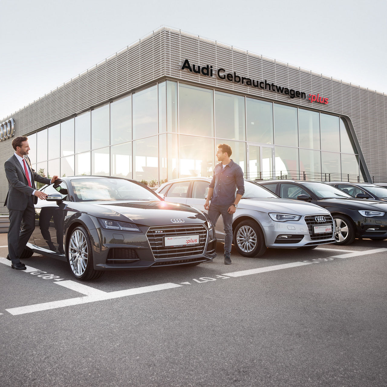 Zwei Männer stehen vor einem Audi TT auf einem Gebrauchtwagenplatz, im Hintergrund ein Gebäude mit der Aufschrift „Audi Gebrauchtwagen :plus“.