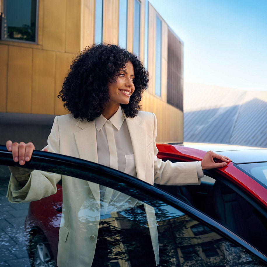 Eine Frau mit lockigem Haar, gekleidet in einem hellen Blazer, steigt aus einem roten Auto aus. Im Hintergrund sind moderne Gebäudefassaden und ein blauer Himmel zu sehen.