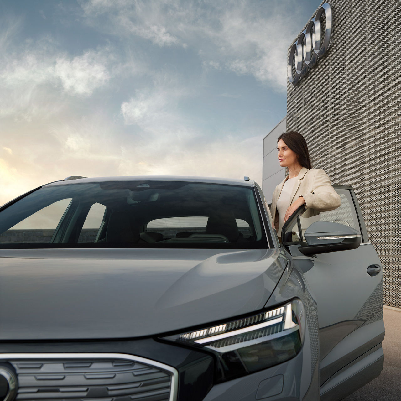 Frau steht an geöffneter Tür eines grauen Audi vor einem Gebäude mit Audi-Logo, der Himmel ist leicht bewölkt.