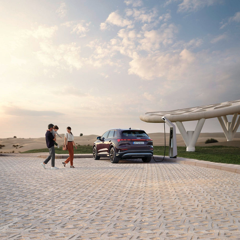 Ein Audi Q4 e-tron lädt an einer futuristischen Ladestation in einer Wüstenlandschaft, während eine Familie mit Kind in der Nähe spaziert.