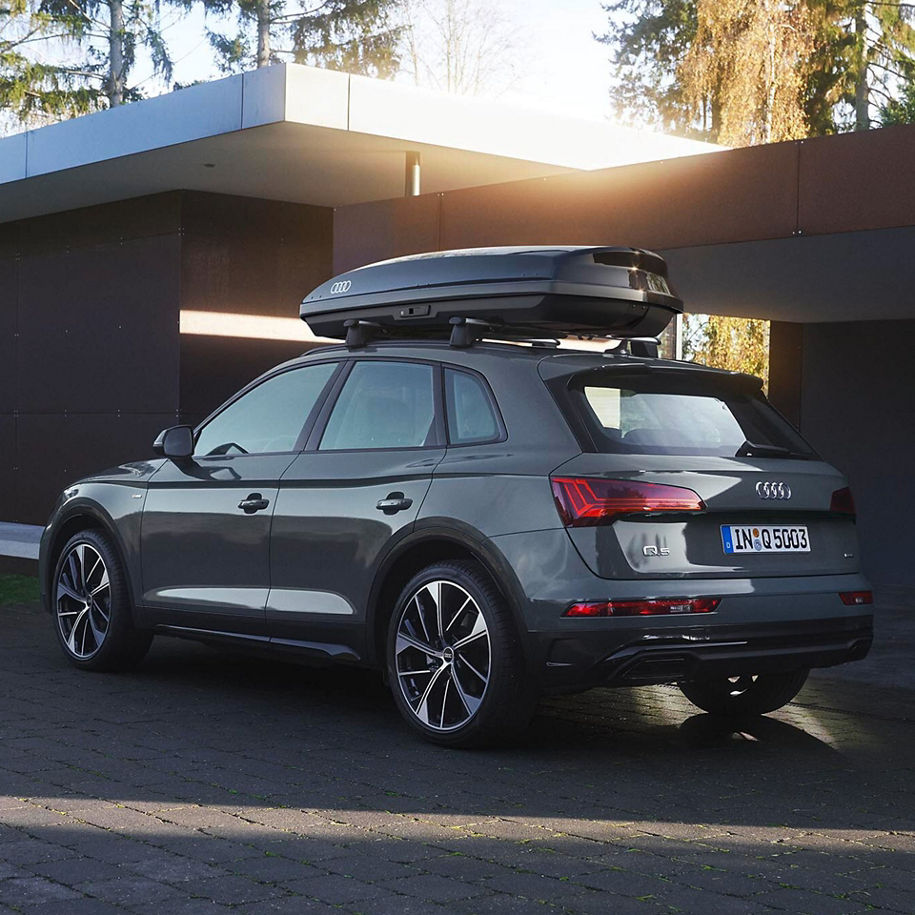 Ein grauer SUV mit einer Dachbox ist auf einer gepflasterten Auffahrt vor einem modernen Gebäude mit minimalistischer Architektur geparkt.