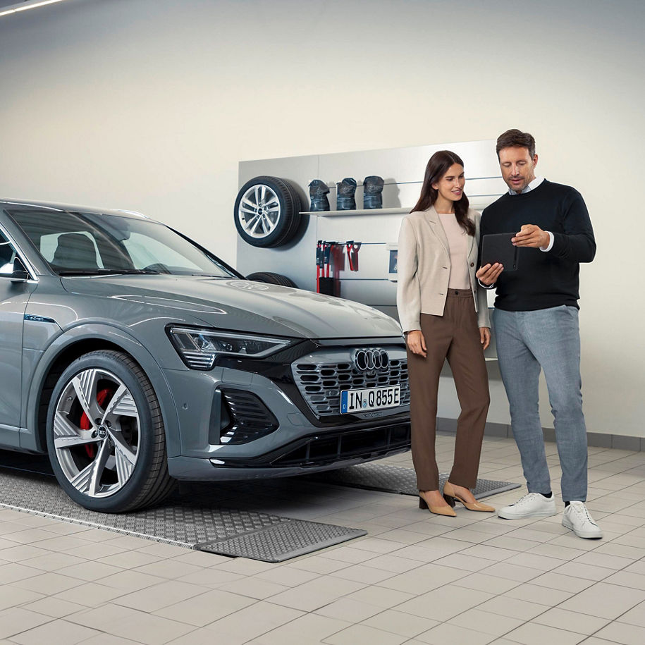 Ein Mann und eine Frau stehen in einer modernen Werkstatt neben einem grauen Audi. Sie betrachten gemeinsam ein Tablet. Im Hintergrund sind Reifen und Werkstattzubehör an der Wand montiert.