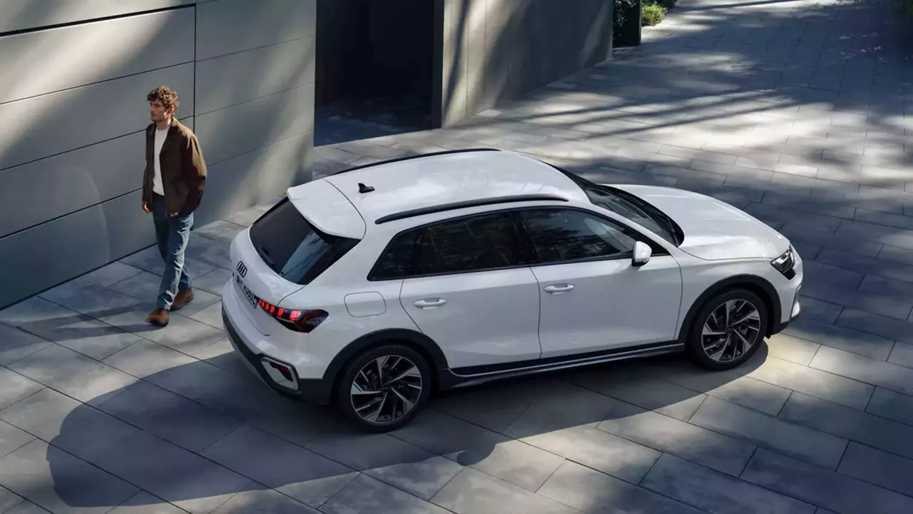 Mann geht an einem weißen Audi SUV auf gepflastertem Hof entlang, modernes Gebäude im Hintergrund, Licht und Schatten auf dem Boden.
