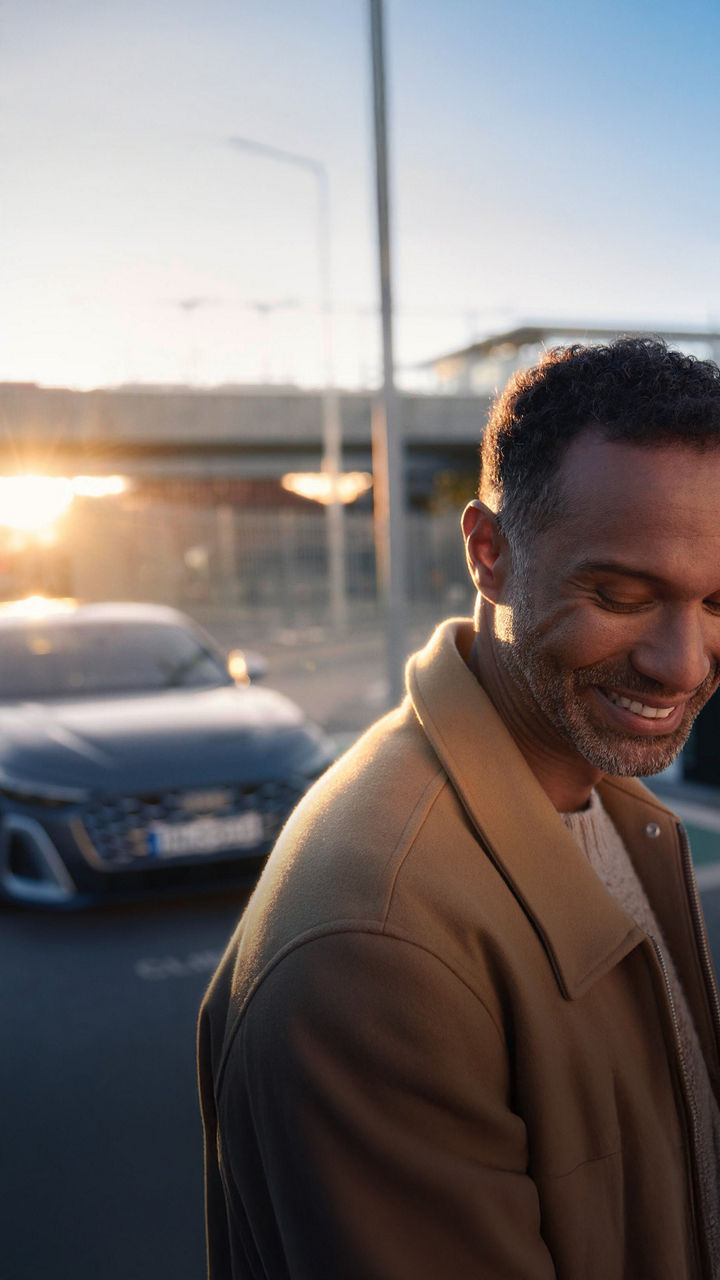 Mann im braunen Mantel steht lächelnd vor einem geparkten Audi bei Sonnenuntergang.