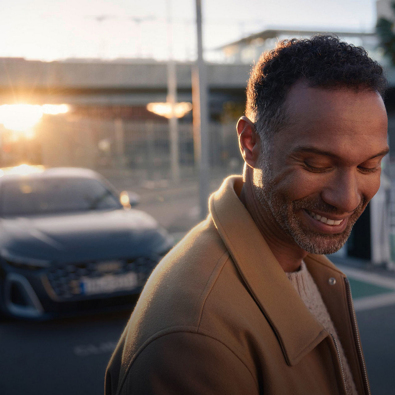 Mann im braunen Mantel steht lächelnd vor einem geparkten Audi bei Sonnenuntergang.