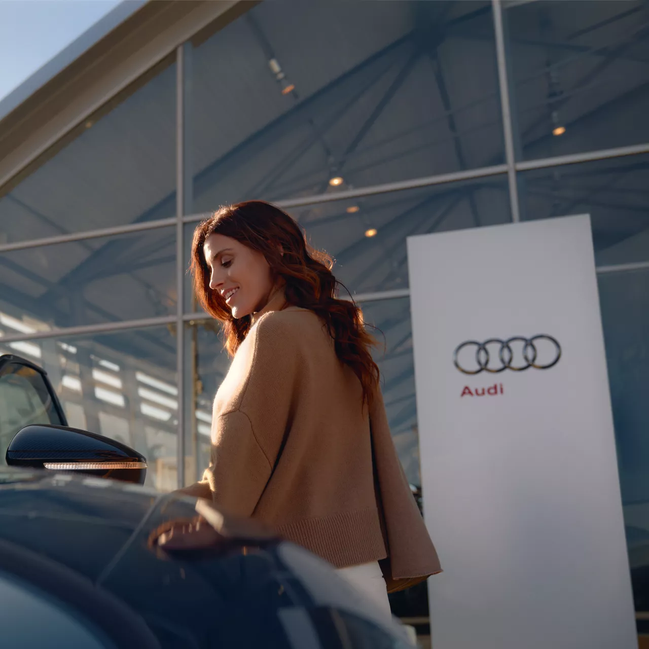 Eine Frau mit langen, kastanienbraunen Haaren steht lächelnd neben einem Audi. Im Hintergrund ist ein modernes Glasgebäude mit einem Audi-Logo und Schriftzug zu sehen.