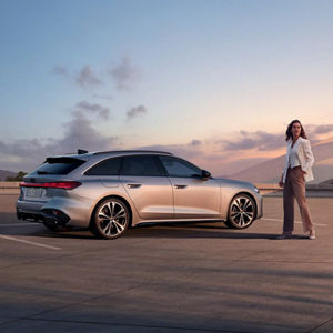 Frau in heller Jacke steht neben einem silbernen Audi auf einem Parkplatz bei Sonnenuntergang.