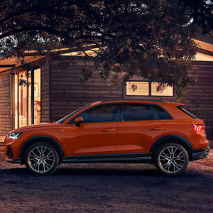 Ein orangefarbener Audi Q3 steht bei Dämmerung vor einem beleuchteten Holzhaus, umgeben von Bäumen und Freizeitgeräten.