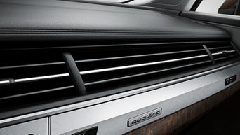 Nahaufnahme eines Lüftungsgitters im Innenraum eines Audi-Fahrzeugs mit „quattro“-Schriftzug auf einer metallischen Blende. Die Materialien wirken hochwertig, mit einer Kombination aus Aluminium, Holzdekor und Leder.