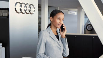 Eine elegant gekleidete Frau im grauen Blazer telefoniert mit einem Smartphone in einem Audi-Autohaus. Im Hintergrund ist das Audi-Logo auf einer metallischen Wand zu sehen, sowie ein Schalter mit der Aufschrift „Teileverkauf“.