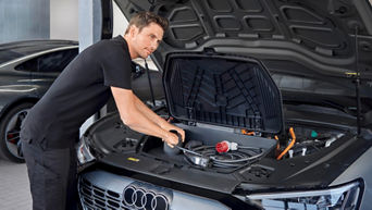 Ein Kfz-Mechatroniker im schwarzen Audi-Polohemd arbeitet am vorderen Stauraum eines Audi E-Autos. Er hantiert mit Ladekabeln und Zubehör, während die Motorhaube geöffnet ist. Im Hintergrund steht ein weiteres Fahrzeug in der Werkstatt.