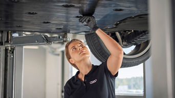 Eine Kfz-Mechatronikerin im schwarzen Audi-Polohemd überprüft die Unterseite eines angehobenen Fahrzeugs in einer Werkstatt. Sie trägt schwarze Handschuhe und inspiziert aufmerksam das Bauteil.