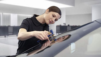 Frau im schwarzen Polo-Shirt repariert konzentriert eine Autoscheibe in heller, moderner Werkstatt.