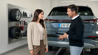Frau im beigefarbenen Blazer und Mann im schwarzen Pullover sprechen in Werkstatt vor grauem Audi Q4, Wand mit Rädern im Hintergrund.