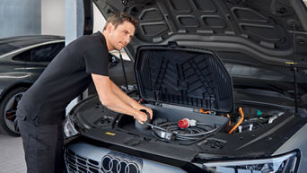 Mann in schwarzer Arbeitskleidung prüft unter geöffneter Motorhaube eines Audi e-tron die Kabel und Anschlüsse im vorderen Stauraum.