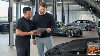 Mechaniker in schwarzer Audi Arbeitskleidung zeigt Kunden auf Tablet etwas, im Hintergrund stehen mehrere Audi Modelle in Werkstatt.