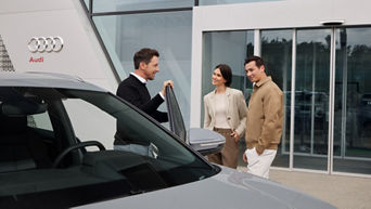 Mann zeigt zwei Personen ein Auto vor Audi Gebäude, sie stehen neben grauem Fahrzeug vor Glasfassade.