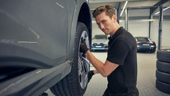 Mechaniker in schwarzer Kleidung befestigt ein Rad an einem grauen Audi auf Hebebühne in der Werkstatt.