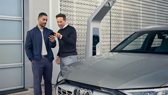 Zwei Männer stehen neben grauem Audi an Ladesäule, einer zeigt dem anderen etwas auf dem Smartphone.