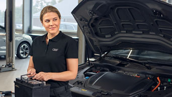 Mechanikerin in schwarzer Audi Arbeitskleidung hält Autobatterie, neben ihr steht ein Auto mit geöffneter Motorhaube in der Werkstatt.