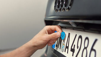 Eine Hand bringt eine blaue Plakette auf einem deutschen Nummernschild an einem schwarzen Audi an.