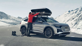 Person in roter Jacke verstaut Skier in Dachbox eines silbernen Audi vor verschneiten Bergen.